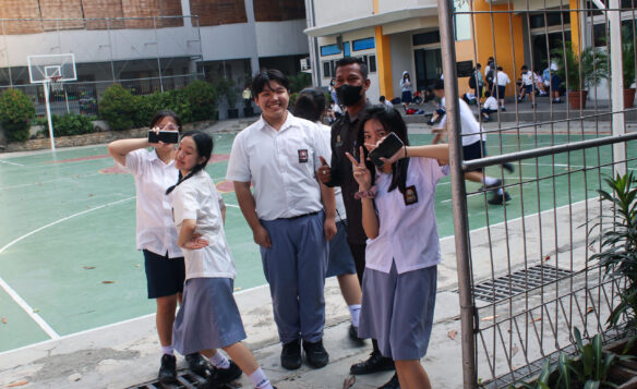 Seorang petugas keamanan berdiri bersama siswa yang sedang bersantai di lingkungan sekolah, menunjukkan keamanan lingkungan pendidikan yang terjaga.